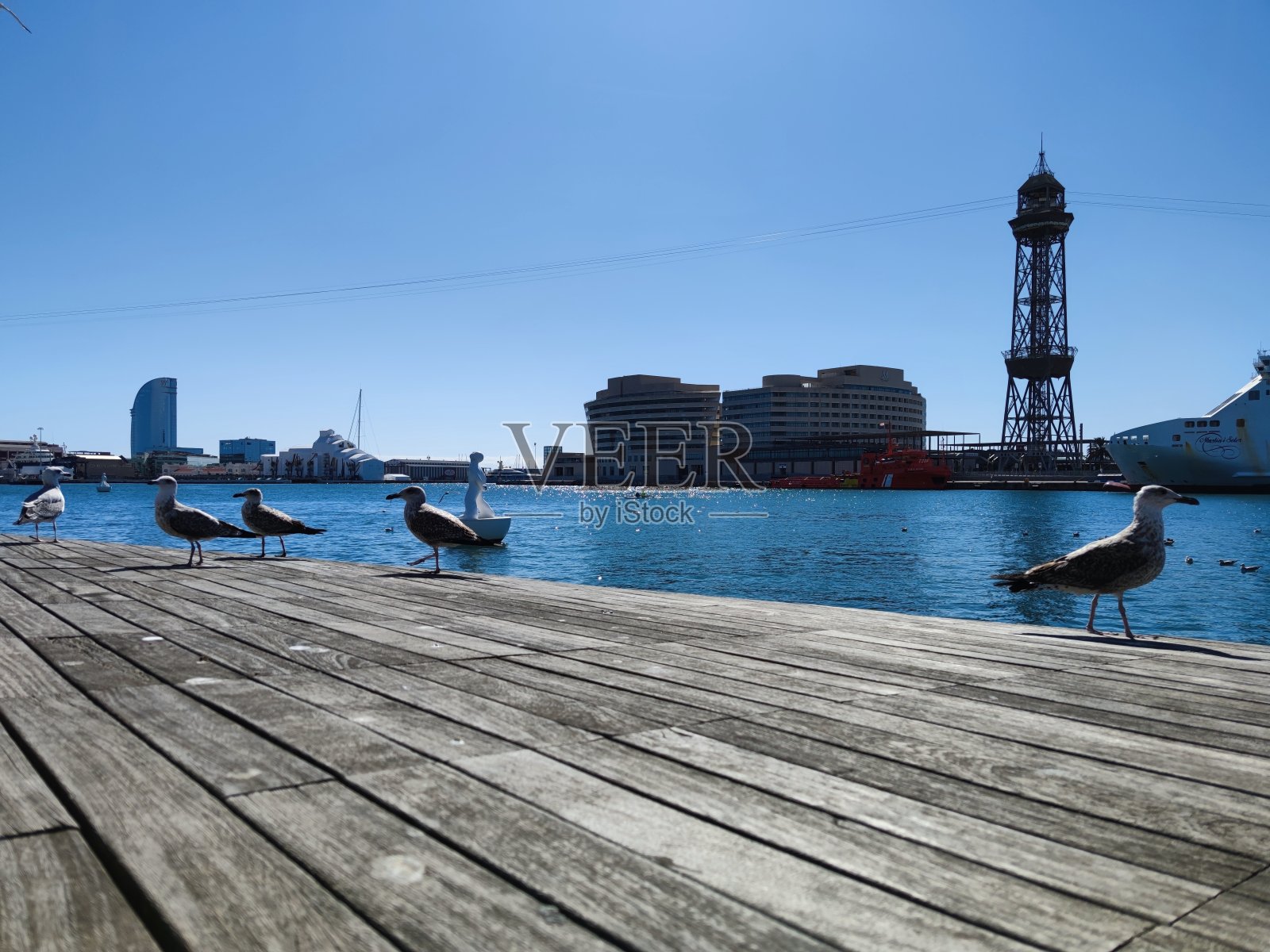 在加泰罗尼亚首府玛丽娜的巴塞罗那Vell港附近的Maremagnum购物中心，海鸥/海鸥在一个波浪木码头的末端的特写，后面是地中海和Vela酒店，W酒店照片摄影图片