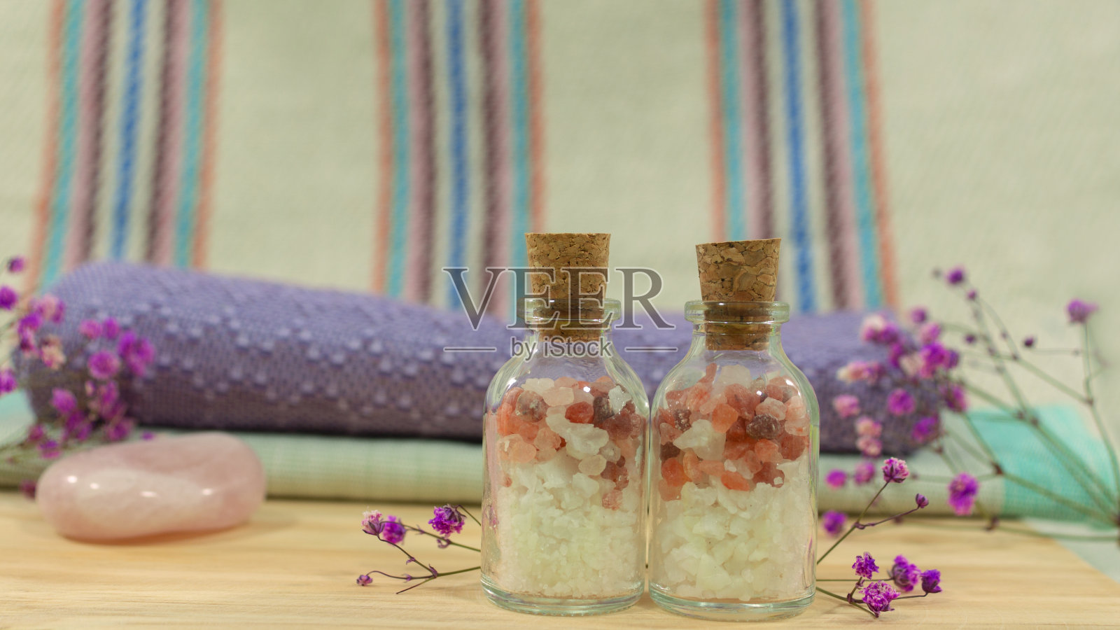 木桌上放着两个装有浴盐的玻璃瓶，背景是毛巾、石头和鲜花照片摄影图片