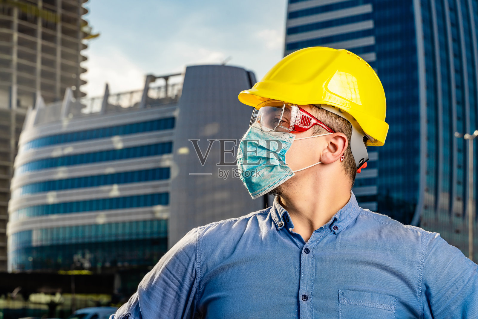 特写的年轻工程师与黄色安全帽和N95防护面具在现代商业建筑和建筑工地前照片摄影图片