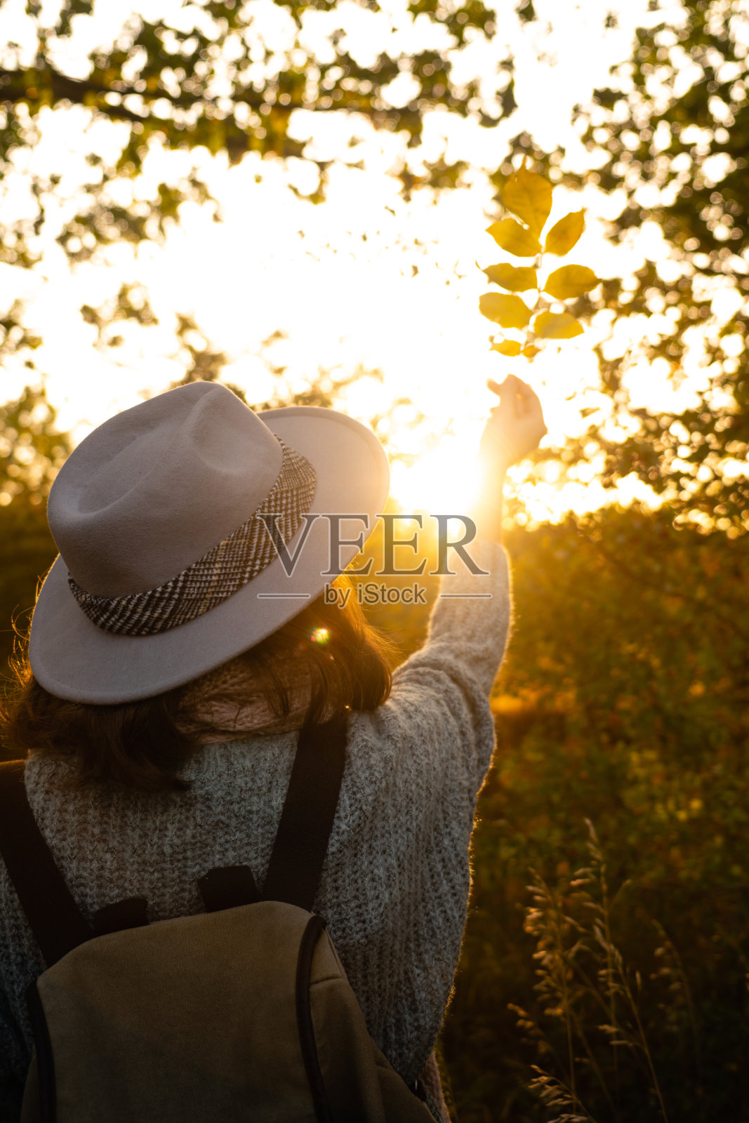 一个戴着帽子，背着背包的女人。日落时，她手里拿着黄叶。照片摄影图片