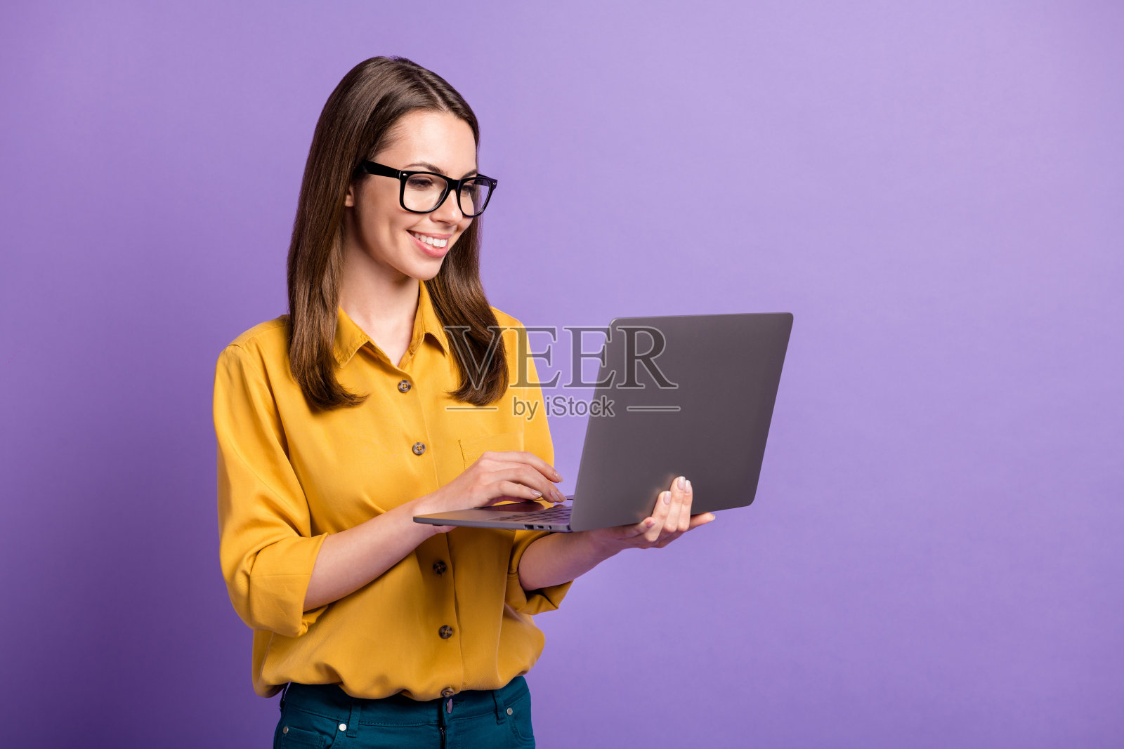 可爱的年轻女孩的照片，拿着电脑看屏幕，戴着眼镜，黄色的衬衫孤立的紫色背景照片摄影图片