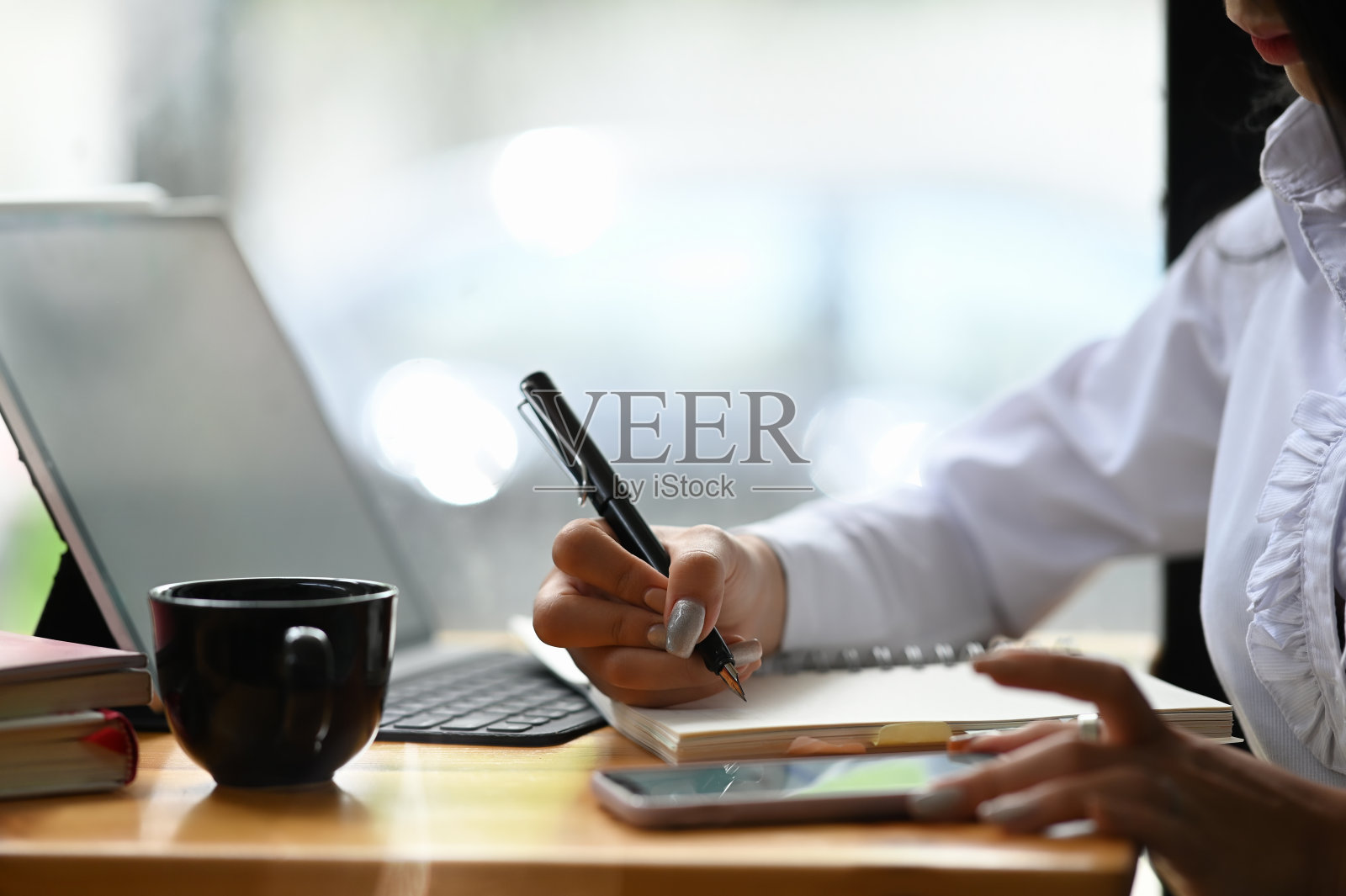 特写女人的手工作在一个智能手机和书写在记事本上与一支笔在办公室。照片摄影图片