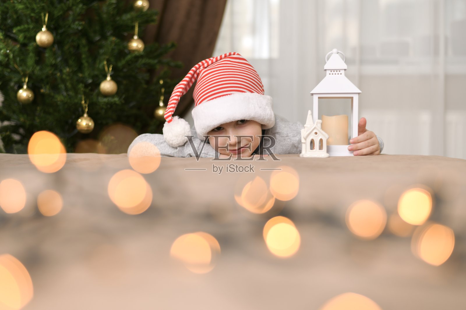 圣诞树旁，一个戴着条纹帽子的孩子躺在他的胳膊上，抱着一盏灯笼和一所房子。照片摄影图片