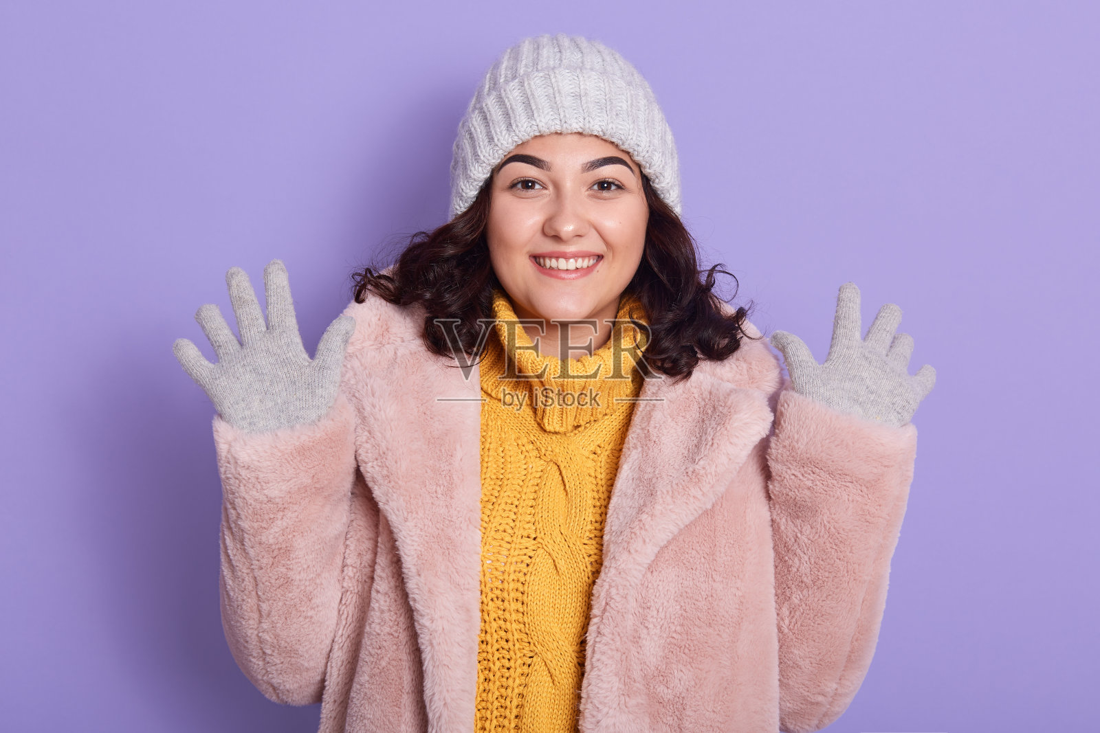 快乐的深色头发的白人妇女在空中伸展双手，有好心情，穿着暖和的外套，毛衣帽和手套，看着镜头露齿微笑，在室内的丁香墙摆姿势。照片摄影图片