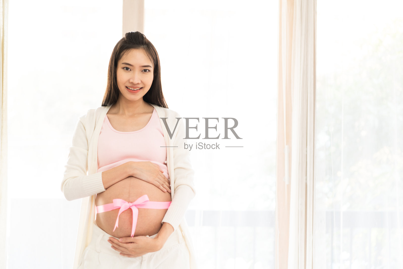 美丽微笑的亚洲孕妇的肖像视图，在她裸露的肚子上有一个大的粉红色丝带。她站在帘子旁边，摸着她的肚子的上半部分。健康的妈妈的概念。照片摄影图片