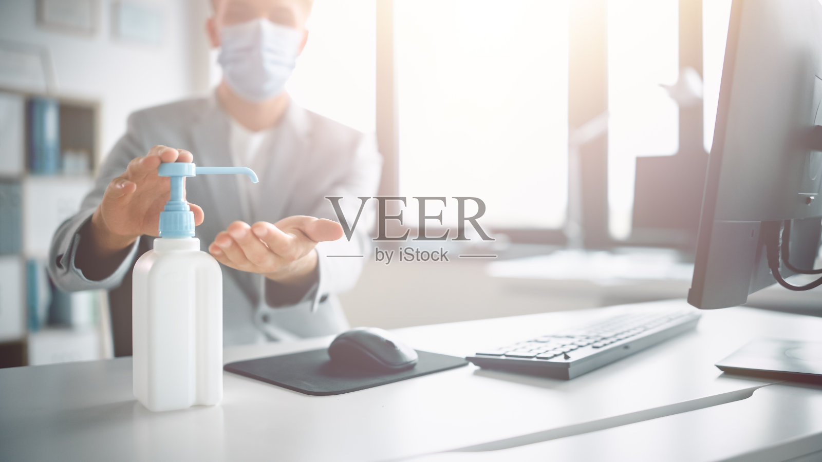 英俊潇洒的年轻人戴着防护面罩，在办公室的电脑前工作时使用洗手液。蓝眼睛金发年轻男性遵循健康和安全指导照片摄影图片