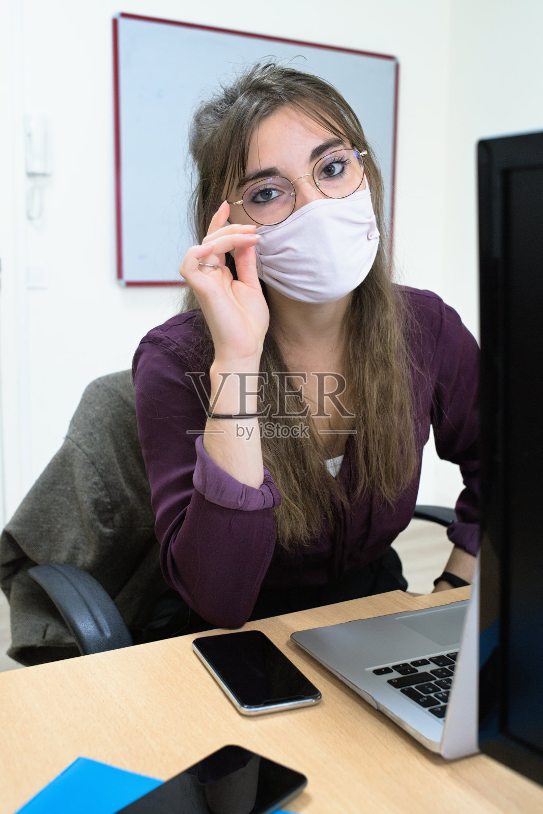 一名年轻女子戴着防冠状病毒的面罩，站在笔记本电脑后面看着相机副本照片摄影图片