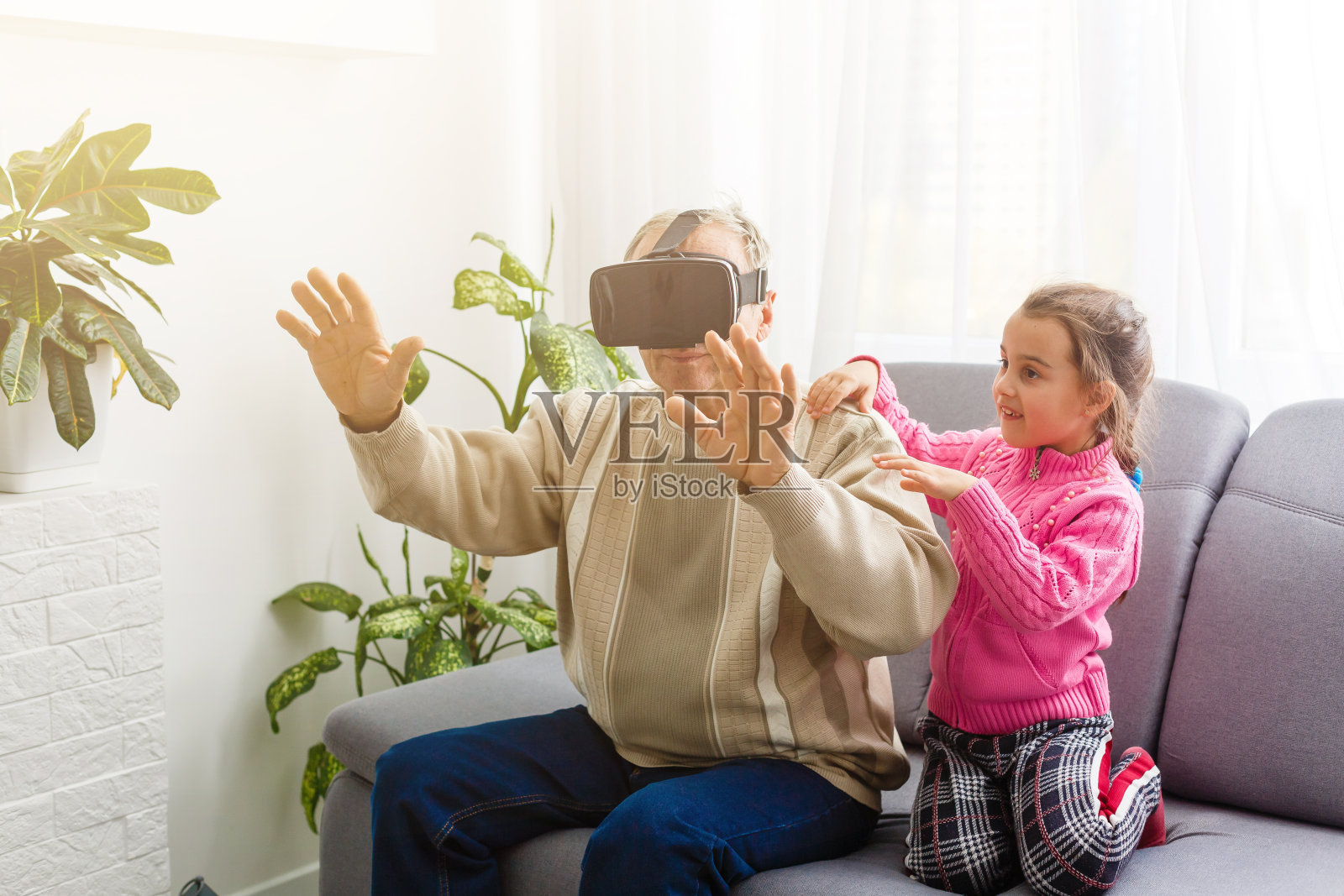 在学校放假期间，孙女和爷爷一起试着演示如何使用VR机。一位戴着VR眼镜坐在家里沙发上大笑的老人照片摄影图片