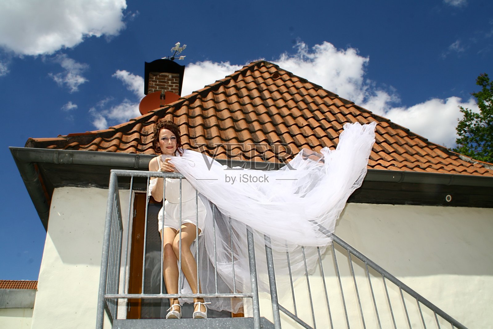 迷人的女人穿着白色的连衣裙和面纱对着白色的墙壁和铁条窗照片摄影图片