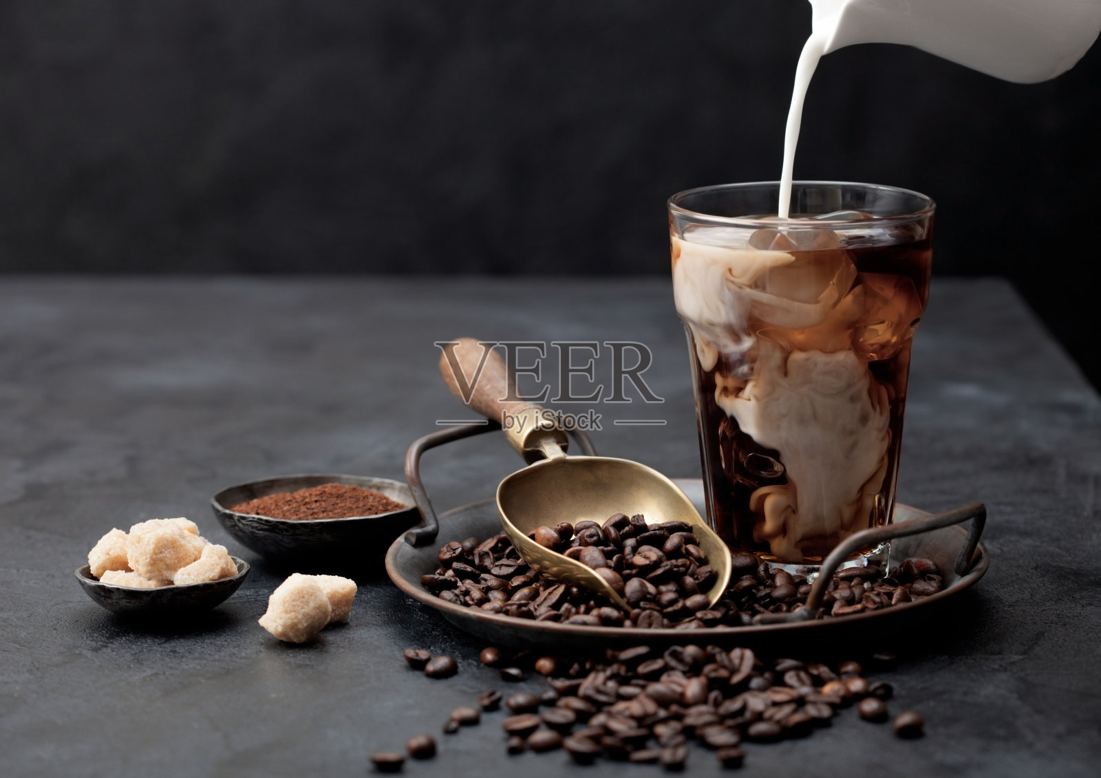 将新鲜牛奶倒入冰黑咖啡托盘上的豆和磨碎的咖啡与蔗糖和黑色的背景。照片摄影图片