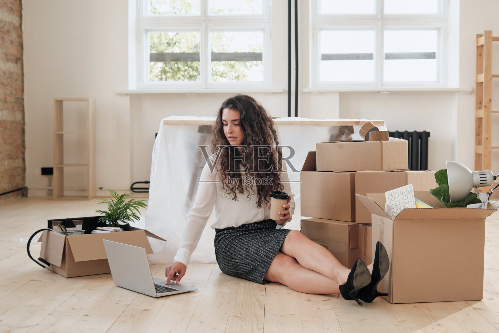 忙碌的年轻秘书穿着正装坐在地板上喝咖啡，而工作与笔记本电脑在空办公室后搬迁照片摄影图片