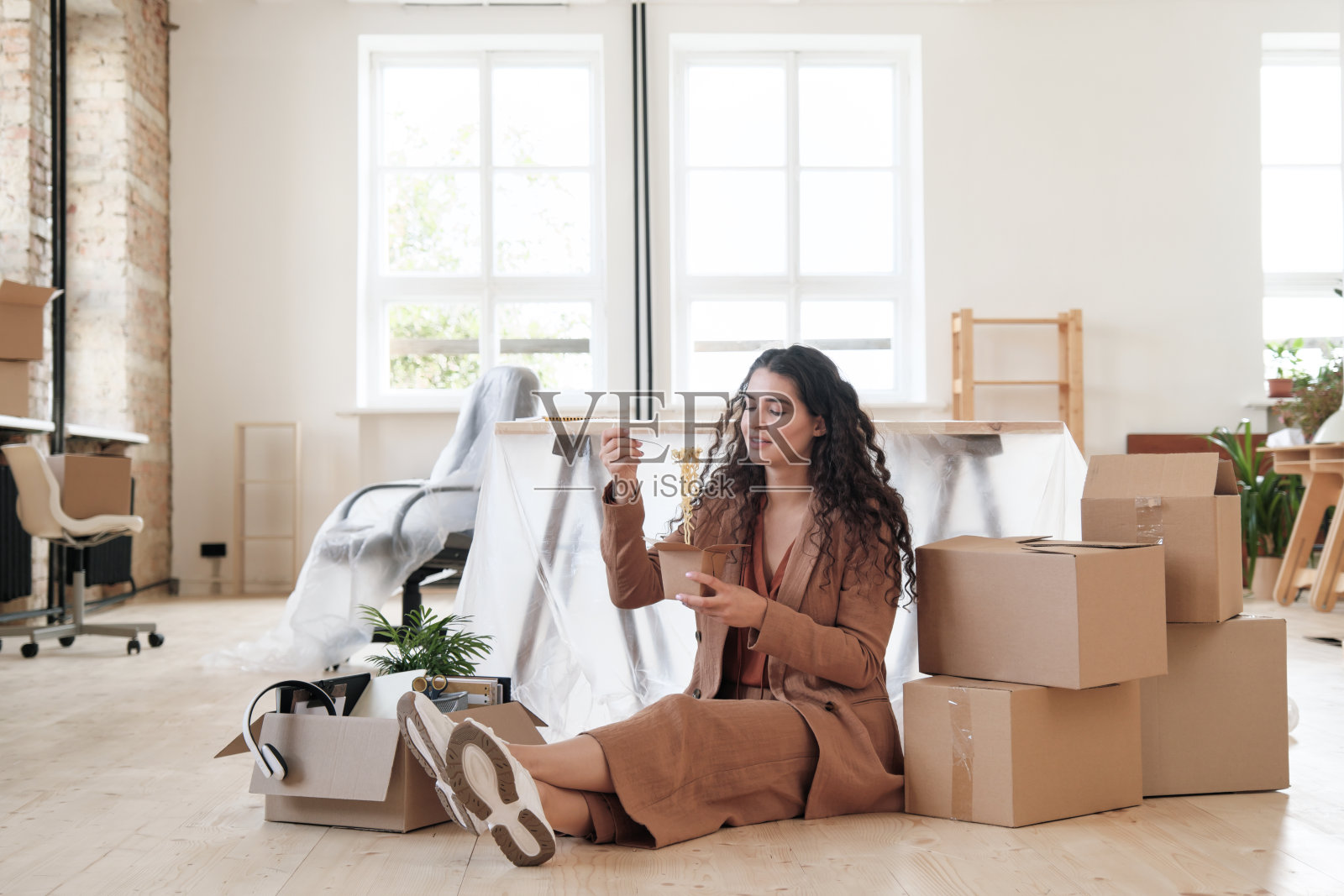 在新的办公室里，无忧无虑的年轻女员工穿着时髦的套装，坐在地板上移动的箱子中间，吃着面条照片摄影图片