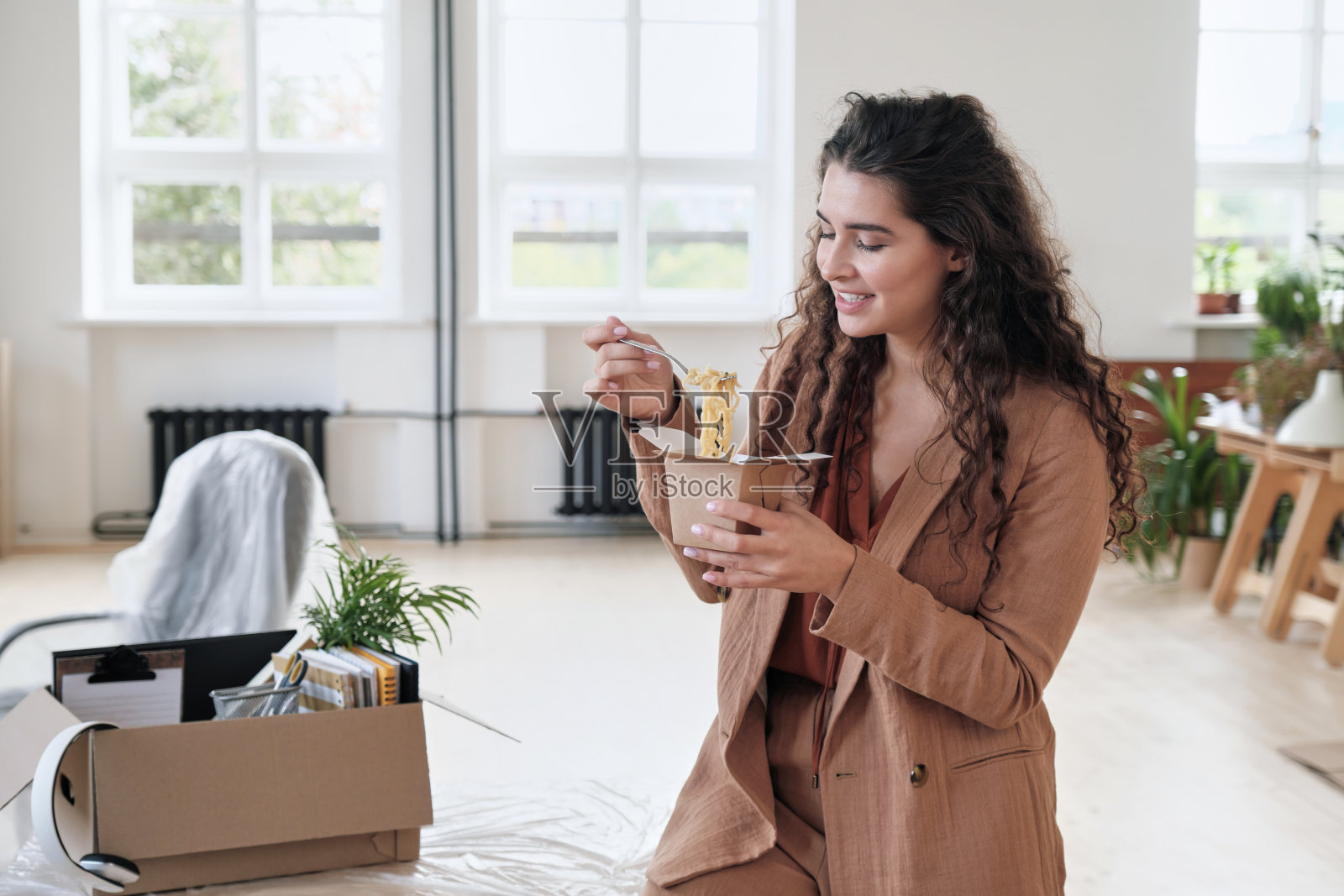 微笑迷人的年轻女商人在夹克靠在桌子移动盒子和吃面条从盒子午休在新办公室照片摄影图片