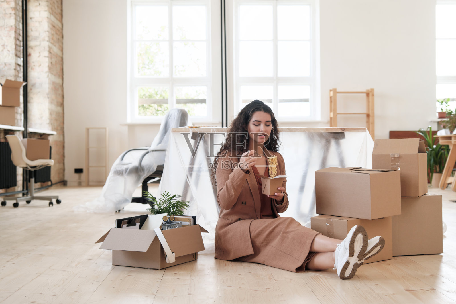 在现代的阁楼办公室里，身着米色套装的年轻办公室女性坐在地板上移动的箱子中间，吃着中国面条照片摄影图片