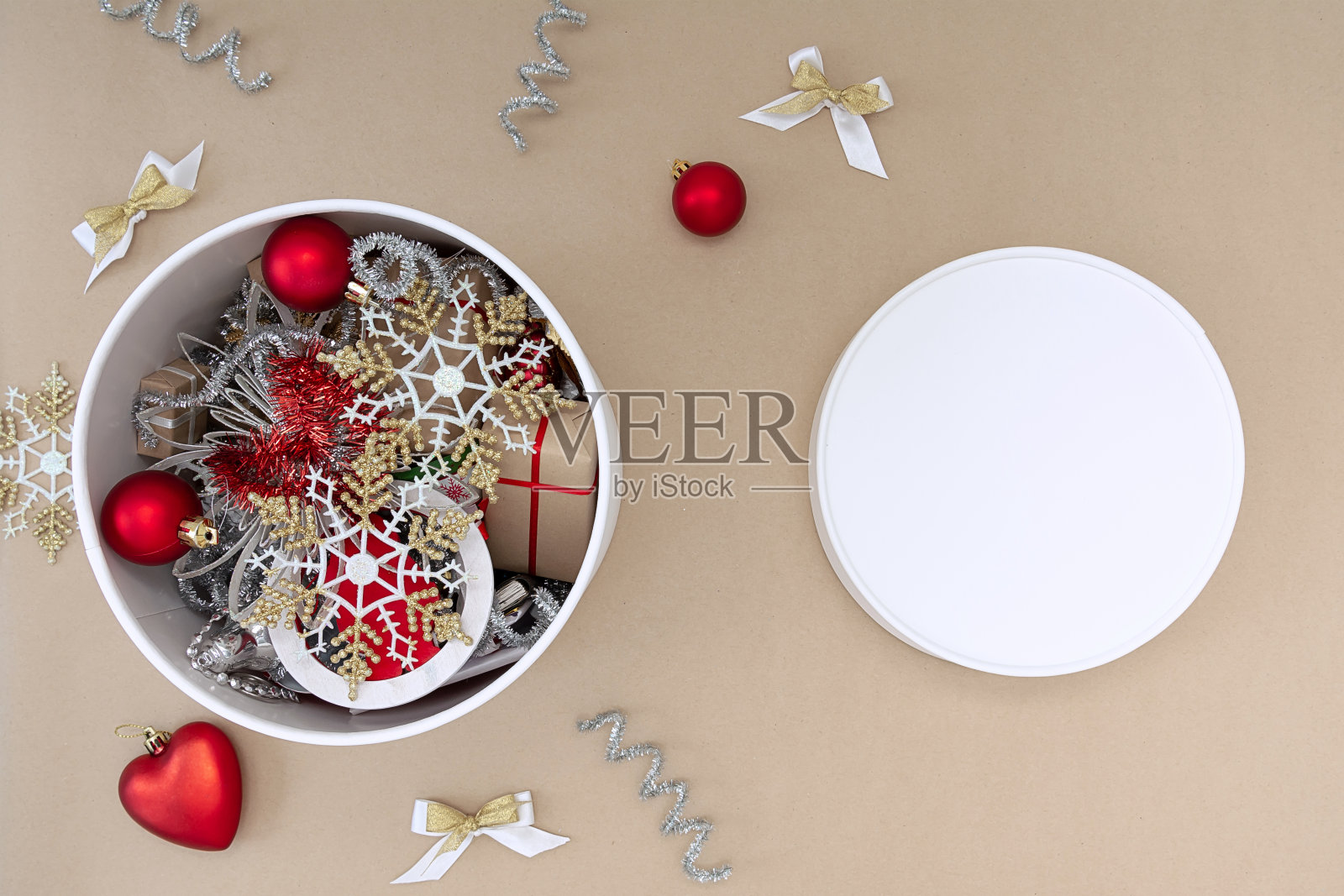 一个白色的圆盒子，里面有圣诞玩具，旁边是米色背景上的红色、白色、金色和银色的装饰品。照片摄影图片