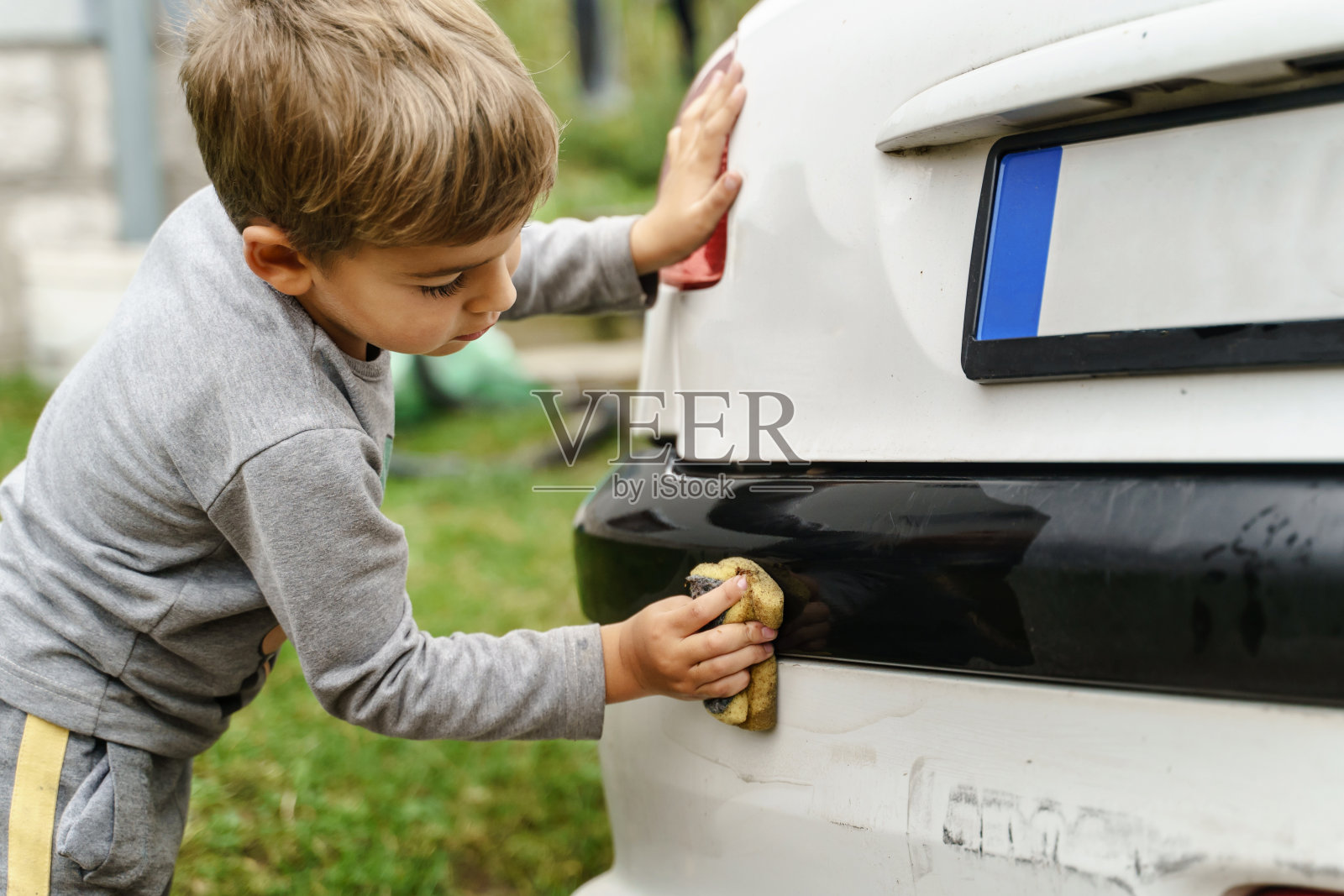 白天，白种人小男孩在室外用海绵洗车——小孩儿在清洗汽车侧视图——小孩儿在摩擦白色车辆的侧视图照片摄影图片