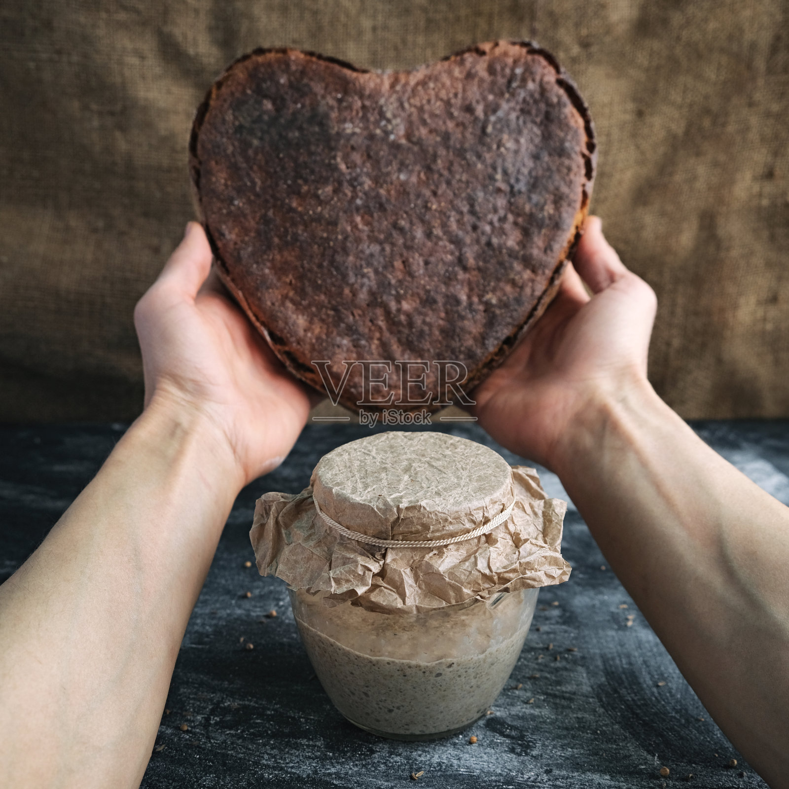 用酸面包和面包师的手拿着心形的黑麦面包的玻璃罐，在棕色的麻布背景上，方形照片摄影图片