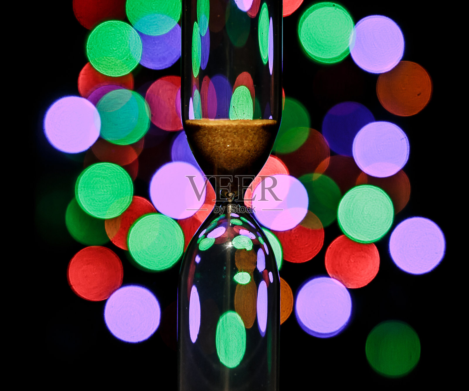 沙漏在中间。沙子洒落在圣诞背景上，散出五颜六色的圣诞彩灯。时间流照片摄影图片