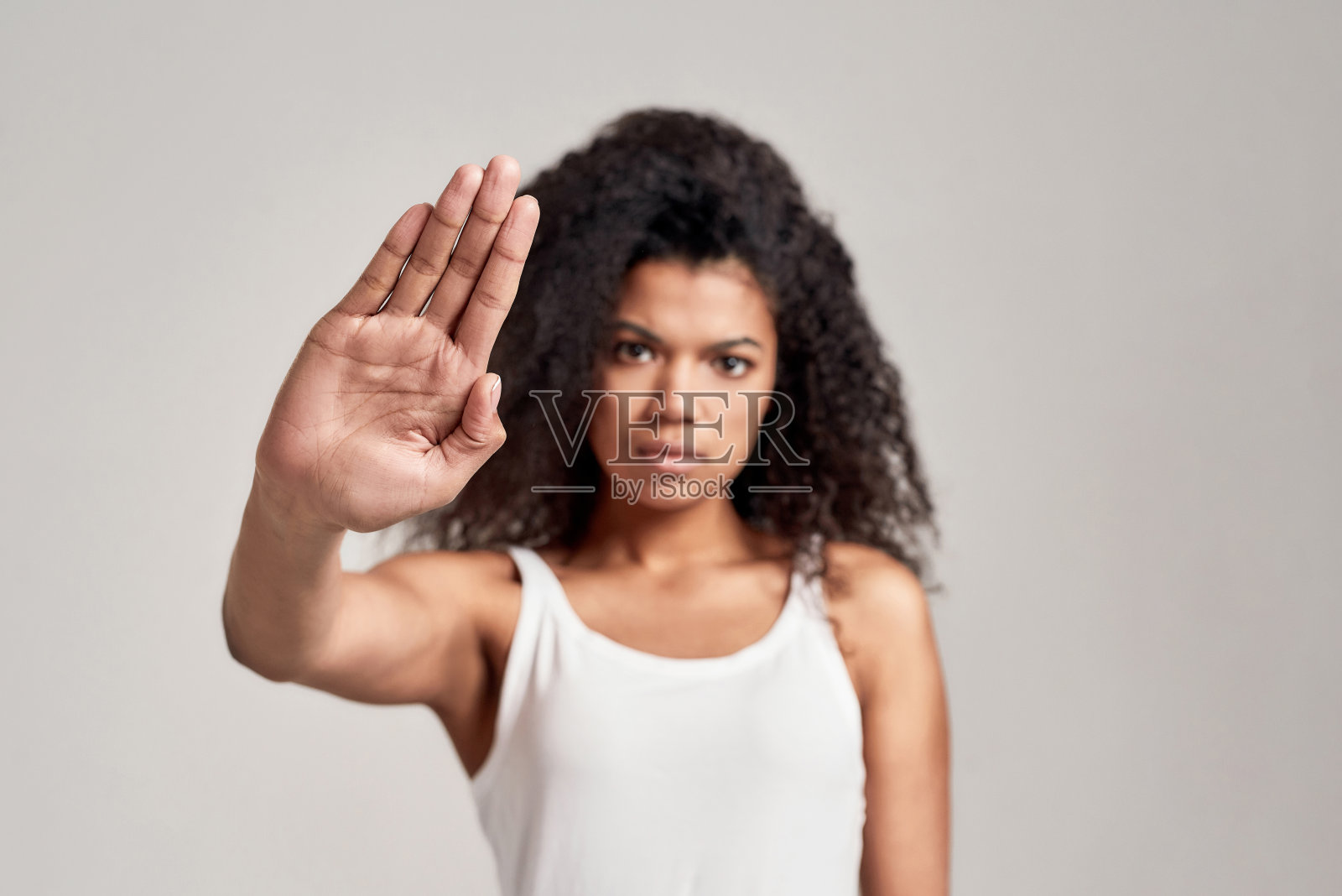 年轻的非洲裔美国妇女的肖像与卷发穿着白色衬衫有严肃自信的样子，而显示，使停止手势孤立在灰色的背景照片摄影图片