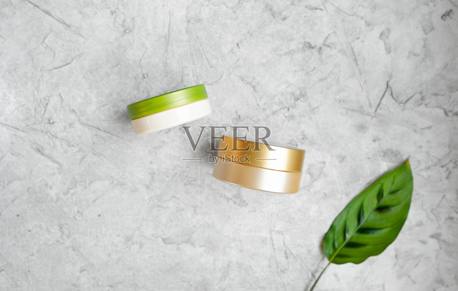 两个金色和绿色的圆形化妆瓶在灰色的背景与绿色植物的叶子，时尚的悬浮化妆布局，选择性的焦点照片摄影图片