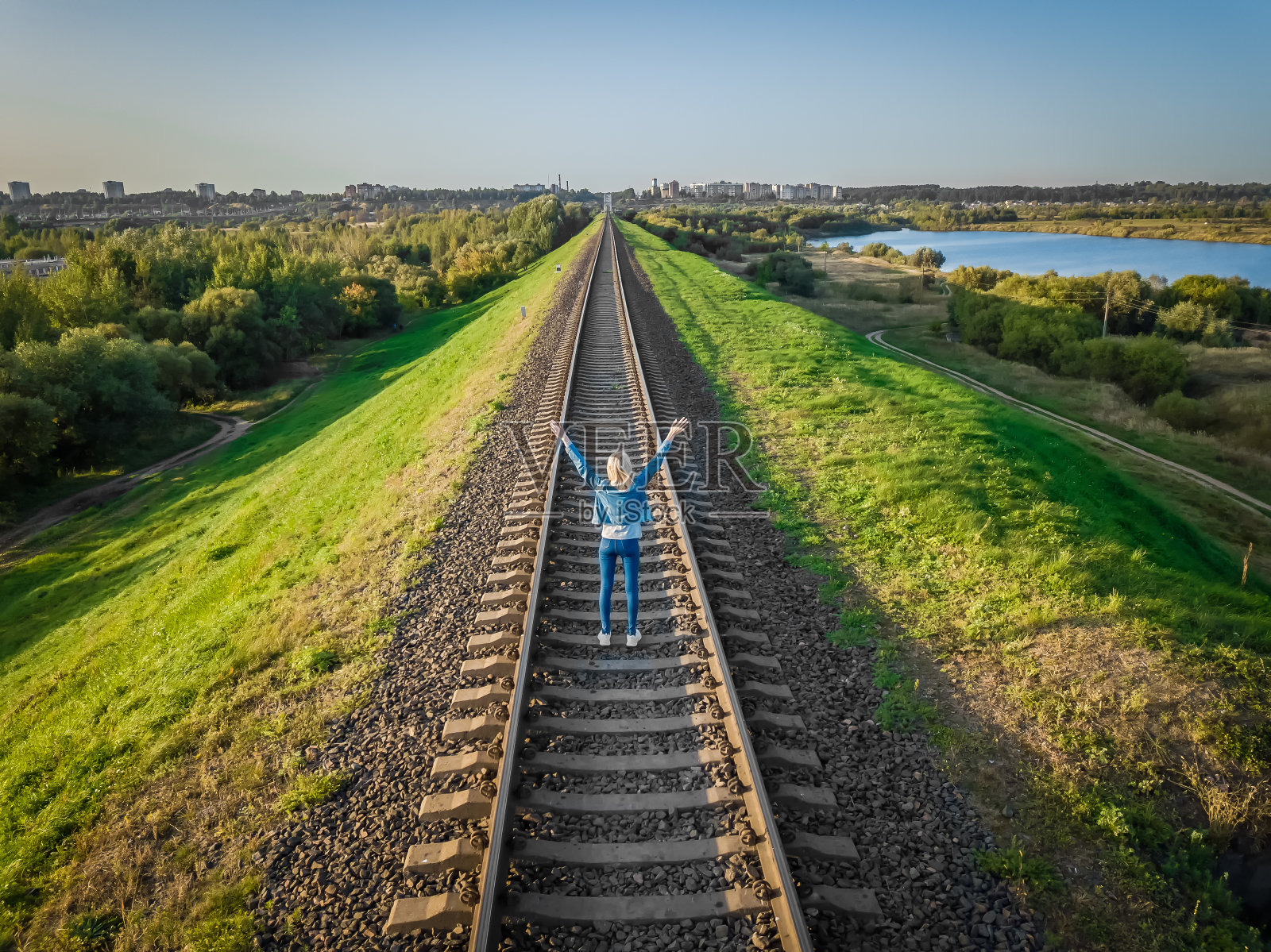 在一个阳光明媚的日子里，从无人机上看到的景象是，穿着牛仔裤和牛仔夹克的金发美女背对着铁轨站着，双手举在空中。观念自由，生活照片摄影图片