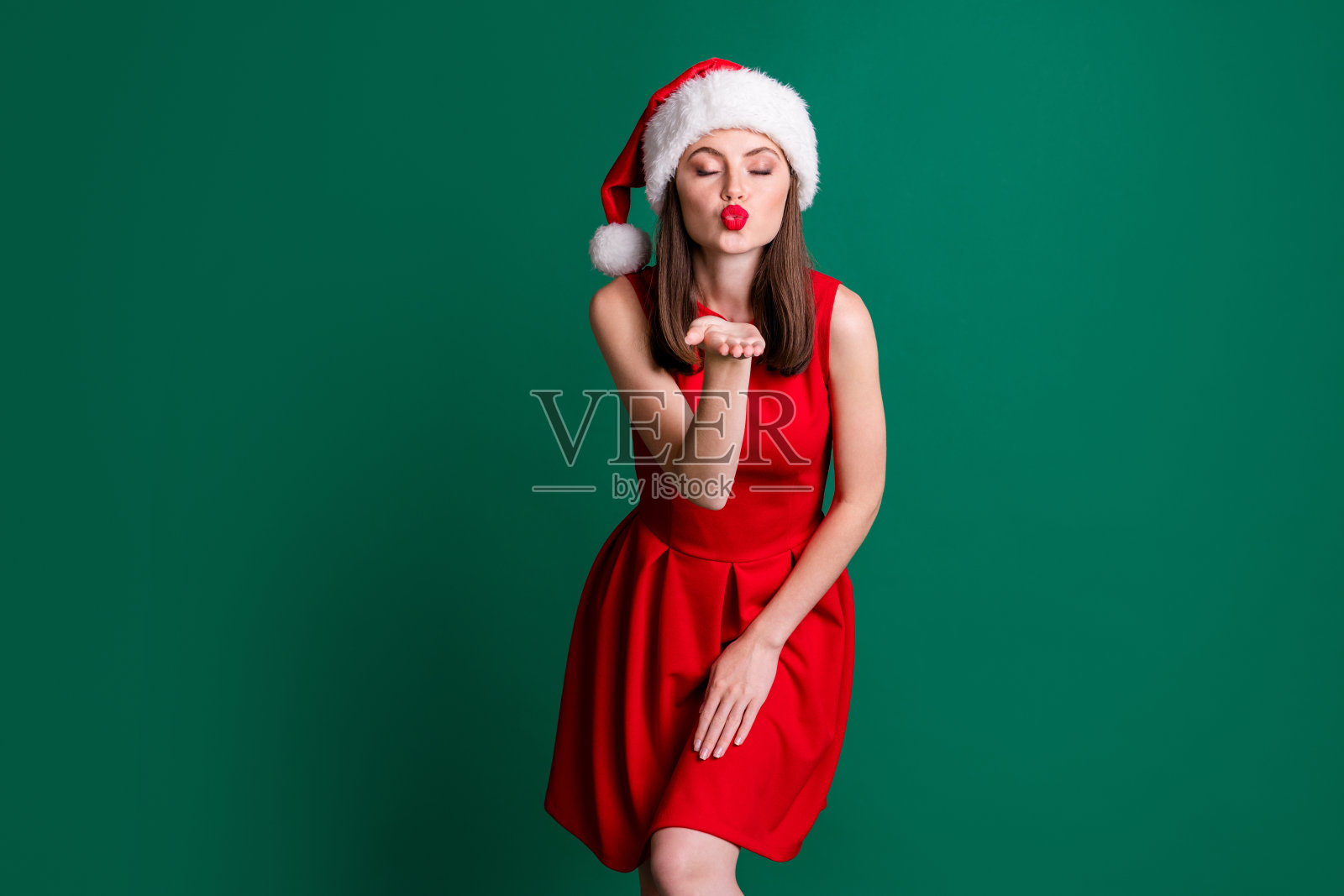 迷人的风骚的女士模特新年派对2021午夜送空中吻在相机上戴圣诞帽红色迷你裙雪女孩服装孤立的绿色柔和的深色背景照片摄影图片