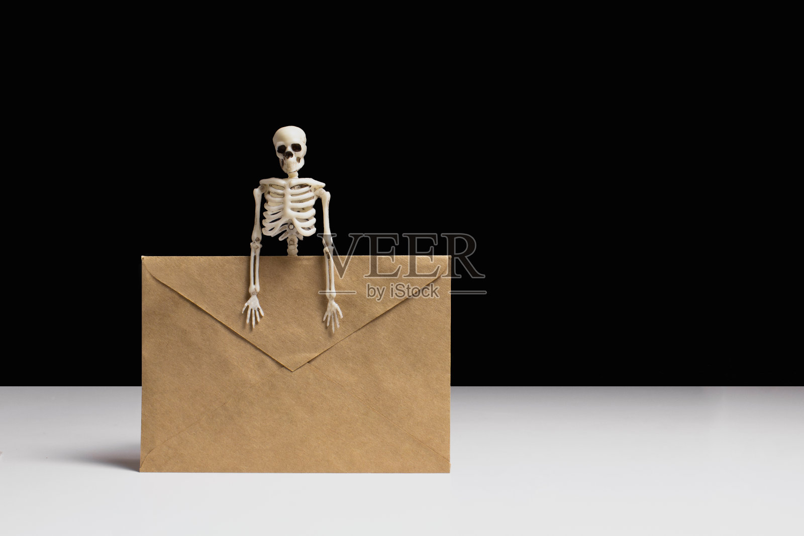 骷髅拿着工艺信封在白色的桌子上，黑色的背景侧视图拷贝空间。万圣节的邀请。节日的庆祝活动。令人毛骨悚然的装饰。照片摄影图片
