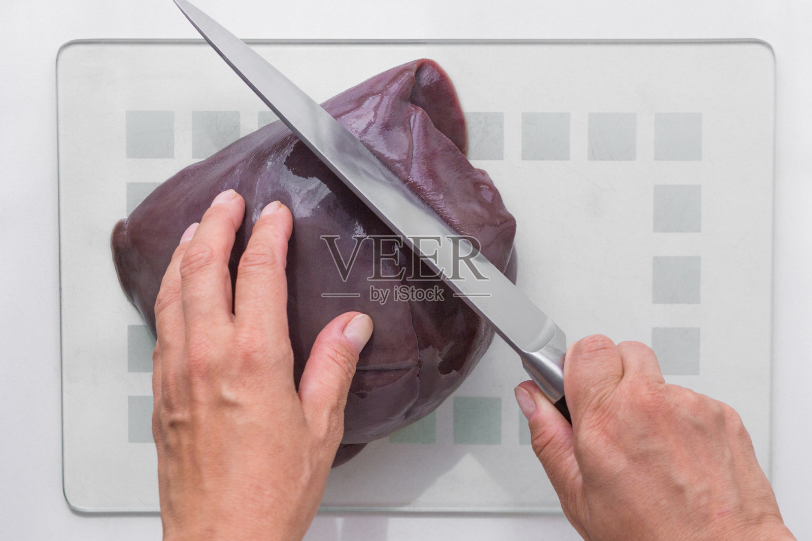 俯视图的妇女手切生块肝脏在玻璃砧板上用刀在白色的背景照片摄影图片