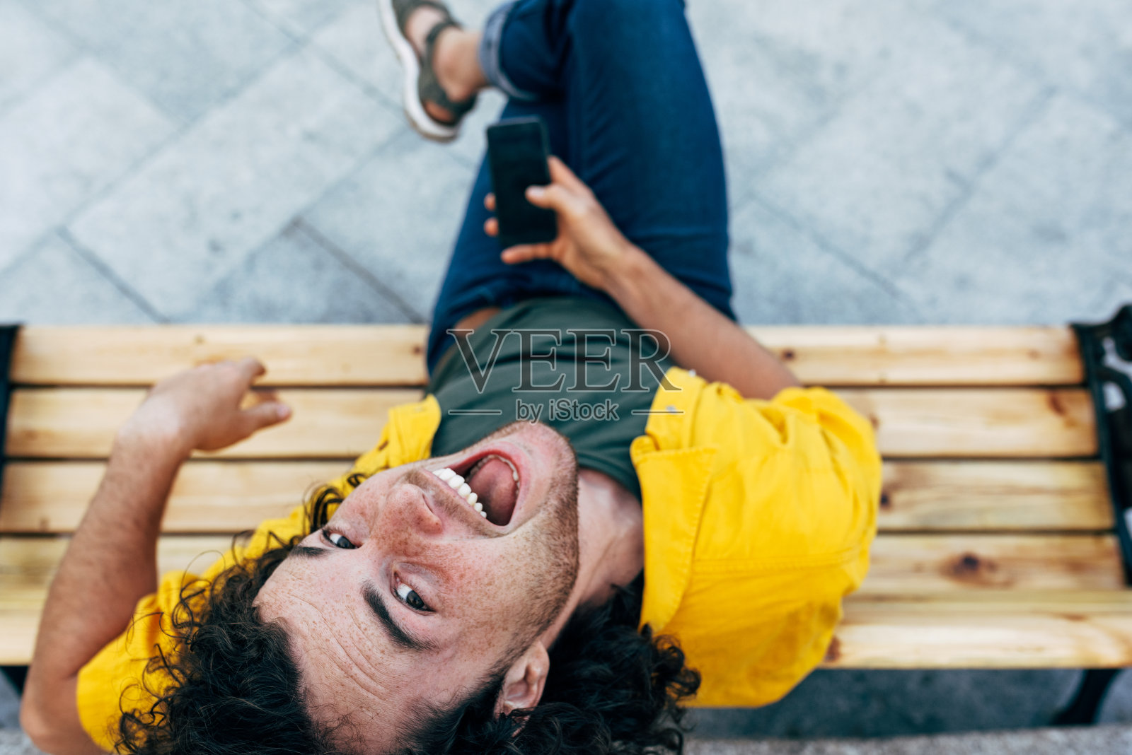 俯视图的年轻人查看手机短信，而坐在户外的长凳上。穿着黄色t恤、绿色t恤和蓝色牛仔裤的快乐男性在城市的街道上通过智能手机在线购买照片摄影图片