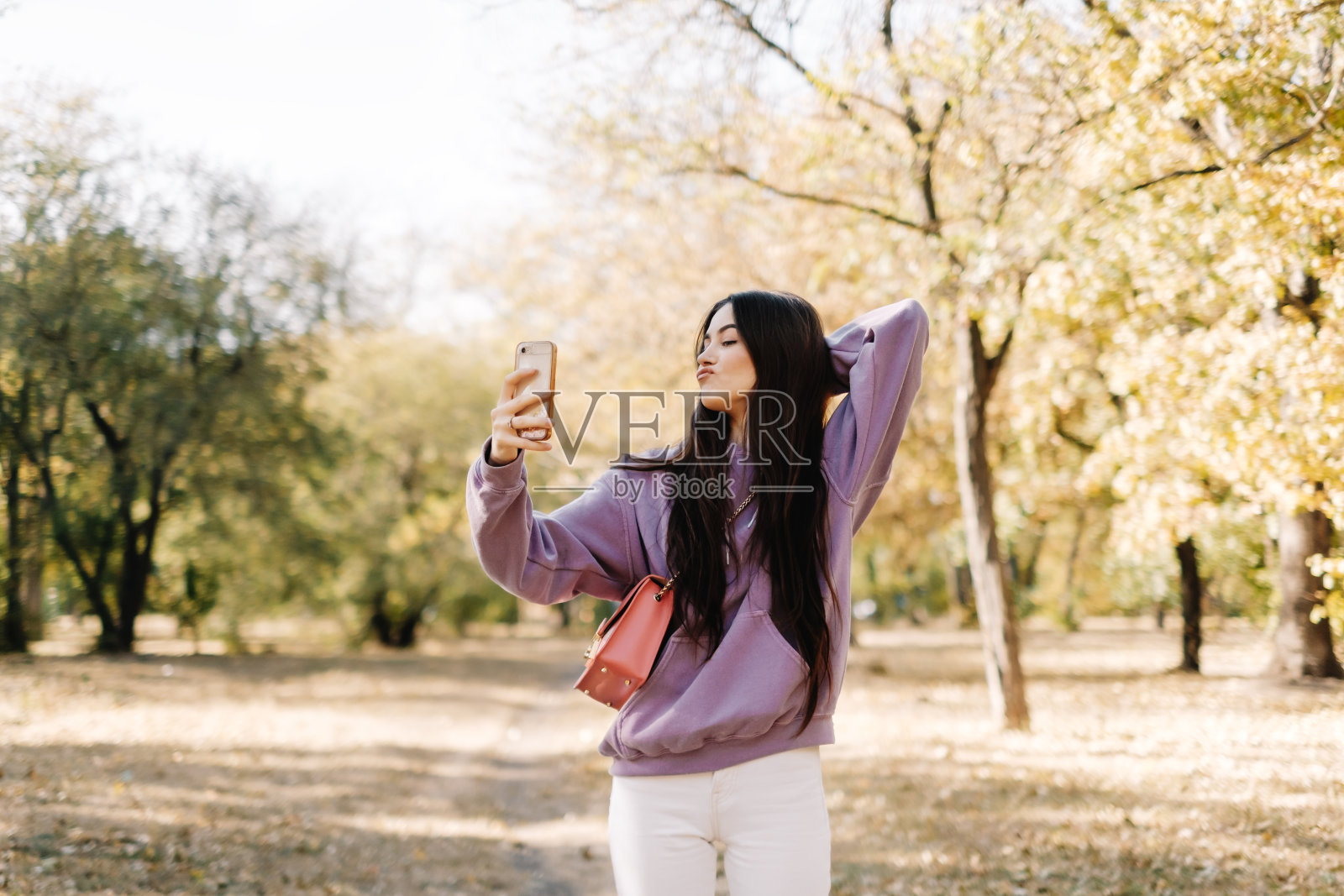 一名女子用智能手机自拍，在公园里享受秋天的天气。照片摄影图片