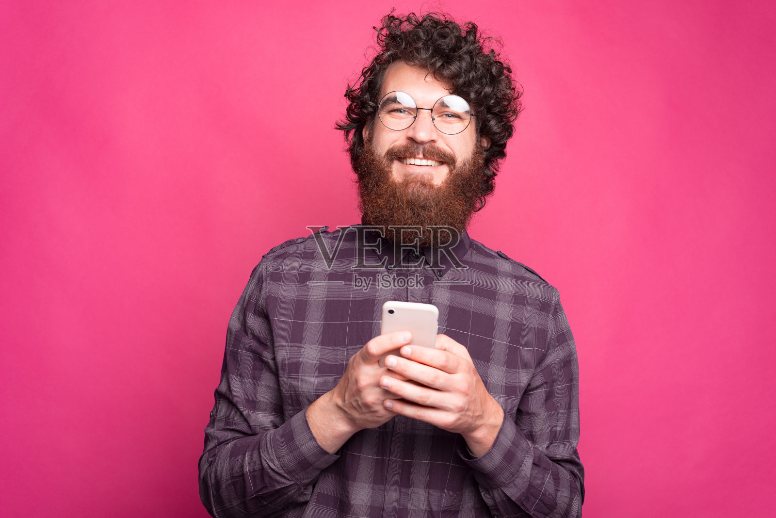 一幅兴高采烈的留着胡子的嬉皮士男子的肖像，他戴着圆框眼镜，拿着智能手机，在镜头前看起来很自信照片摄影图片