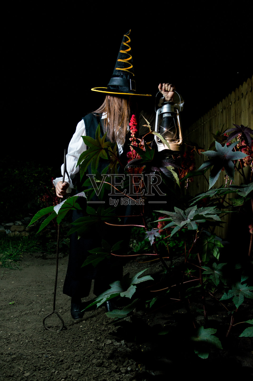 一个女人戴着黑色的锥形帽子，穿着黑色的万圣节服装，手里拿着一盏灯，晚上在花园里有一个金属钩子。照片摄影图片