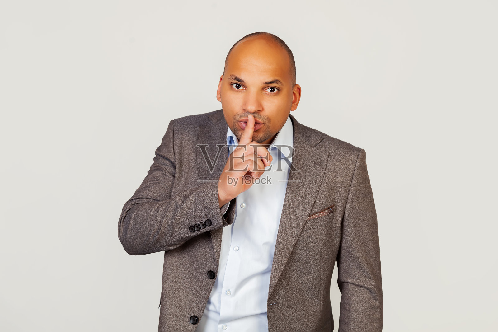 一位年轻的非裔美国商人，穿着夹克，把食指压在嘴唇上，要求不要讲故事，闭上嘴，不要在错误的时刻开玩笑。沉默的迹象。照片摄影图片