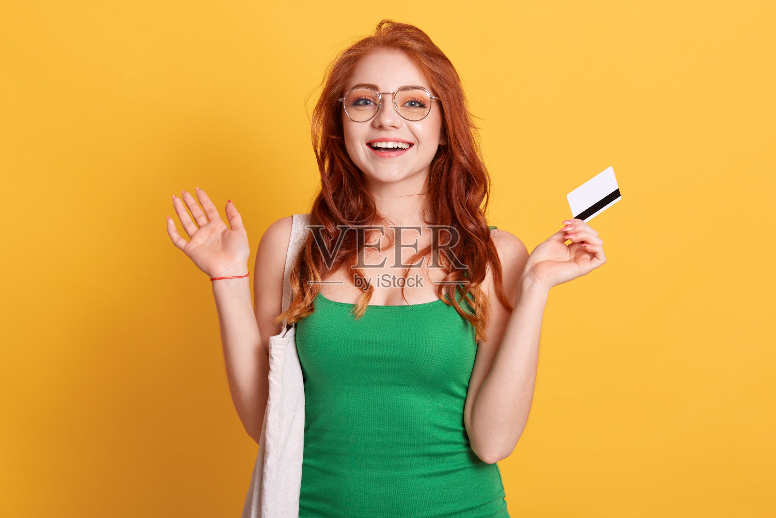 一名年轻的白人女子拿着购物袋和空白的信用卡，双手张开，表情激动，穿着绿色的t恤，有红色的卷发。照片摄影图片