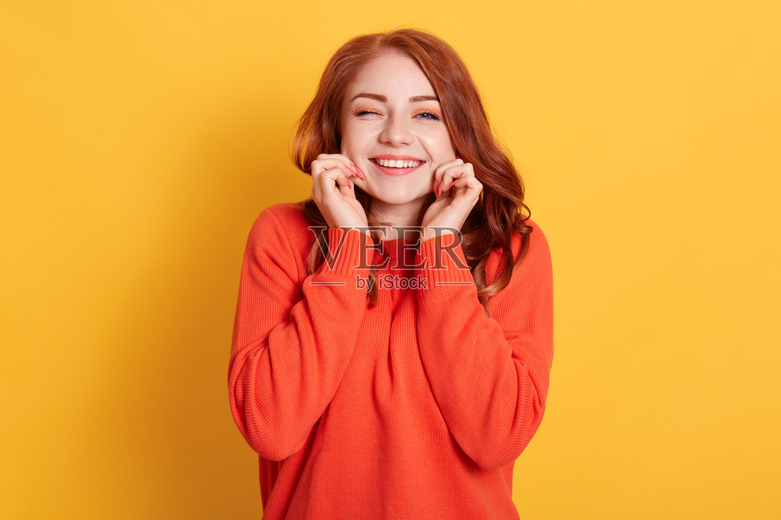 微笑快乐美丽的女士捏她的脸颊，看着微笑直接对着相机和眨眼，穿着橙色毛衣，站在黄色的背景。照片摄影图片