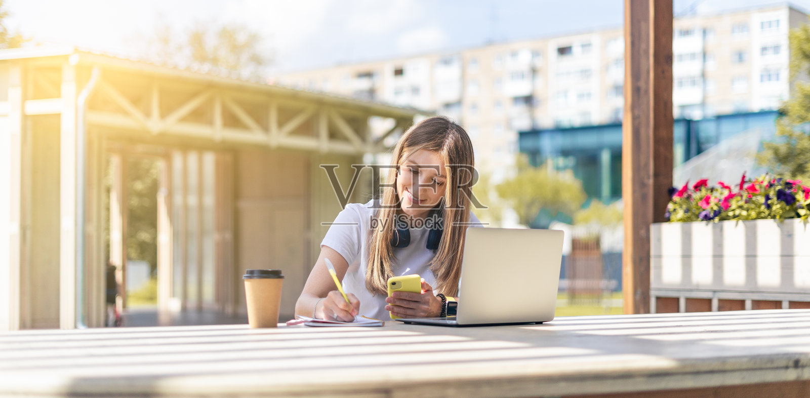 戴着耳机的女人在笔记本上写字，在公园里用笔记本。快乐微笑的女孩一边工作一边喝咖啡。使用电脑。远程学习、在线教育和网上商店照片摄影图片