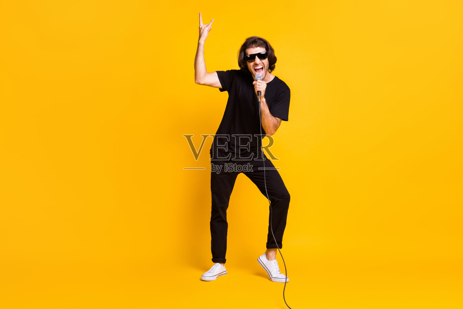 全身照的年轻人拿着麦克风唱张嘴显示角穿黑色t恤裤子白色运动鞋太阳镜孤立黄色背景照片摄影图片