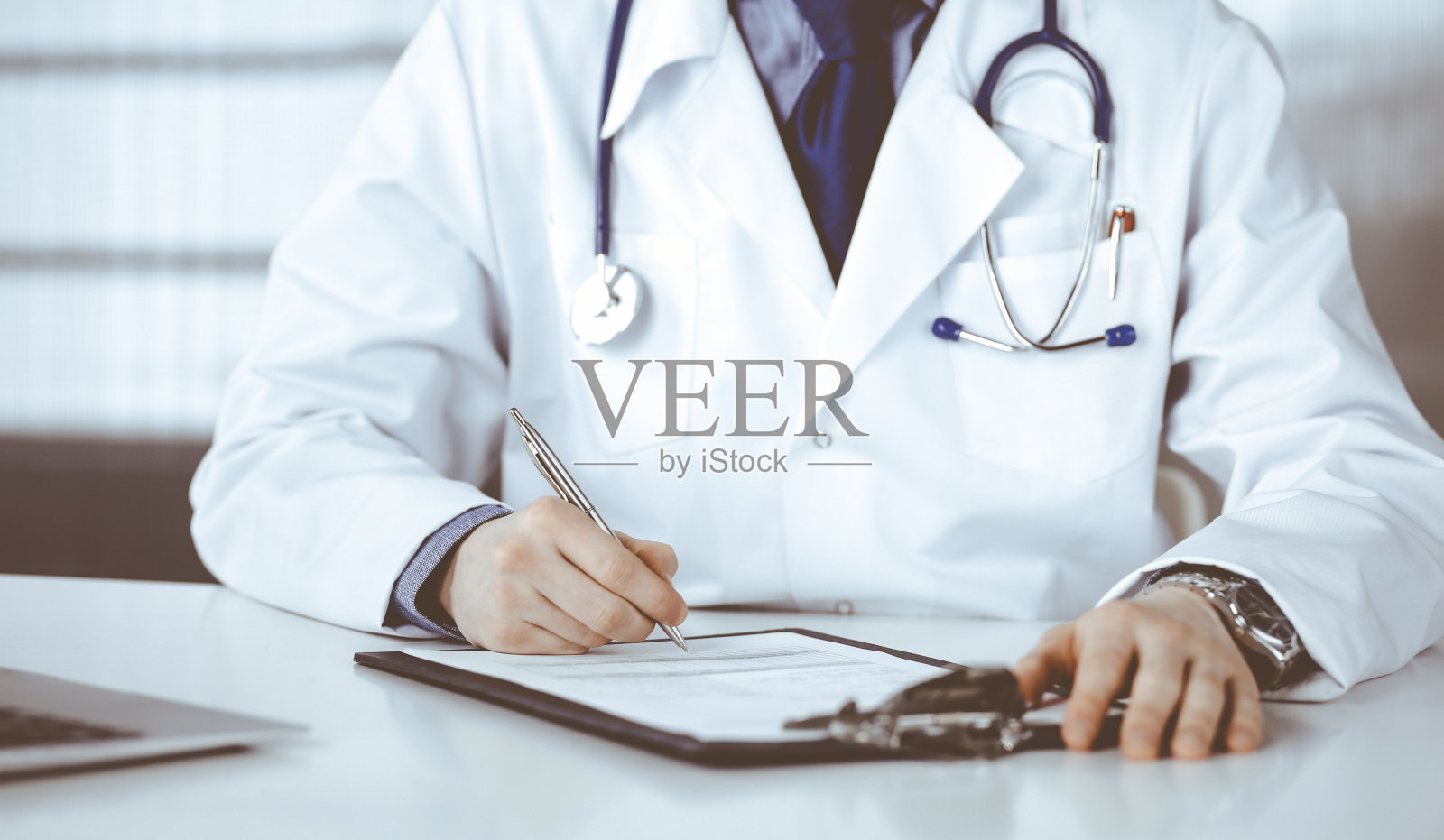 不知名的男医生坐在他的工作地点的诊所和工作与剪贴板，特写。年轻医生在工作。完善的医疗服务，医学理念照片摄影图片