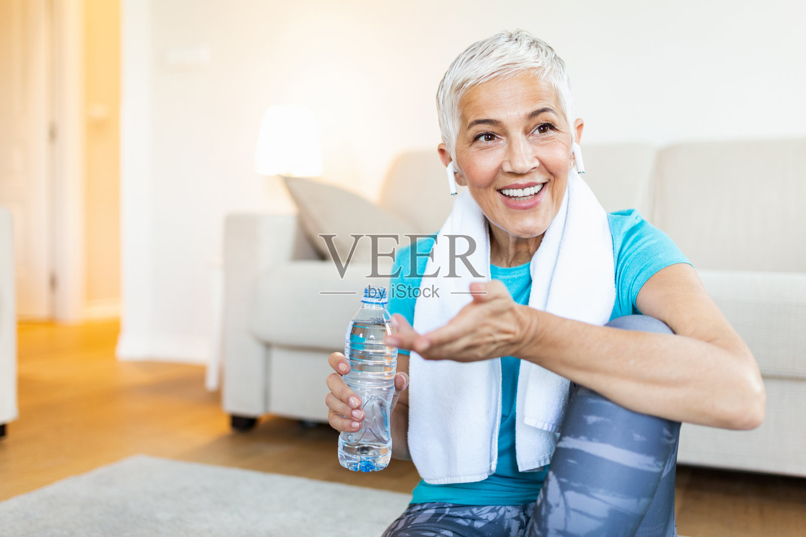 锻炼后戴耳机的老年妇女。拿着一瓶水。成熟的健壮女性在一次良好的锻炼后休息照片摄影图片