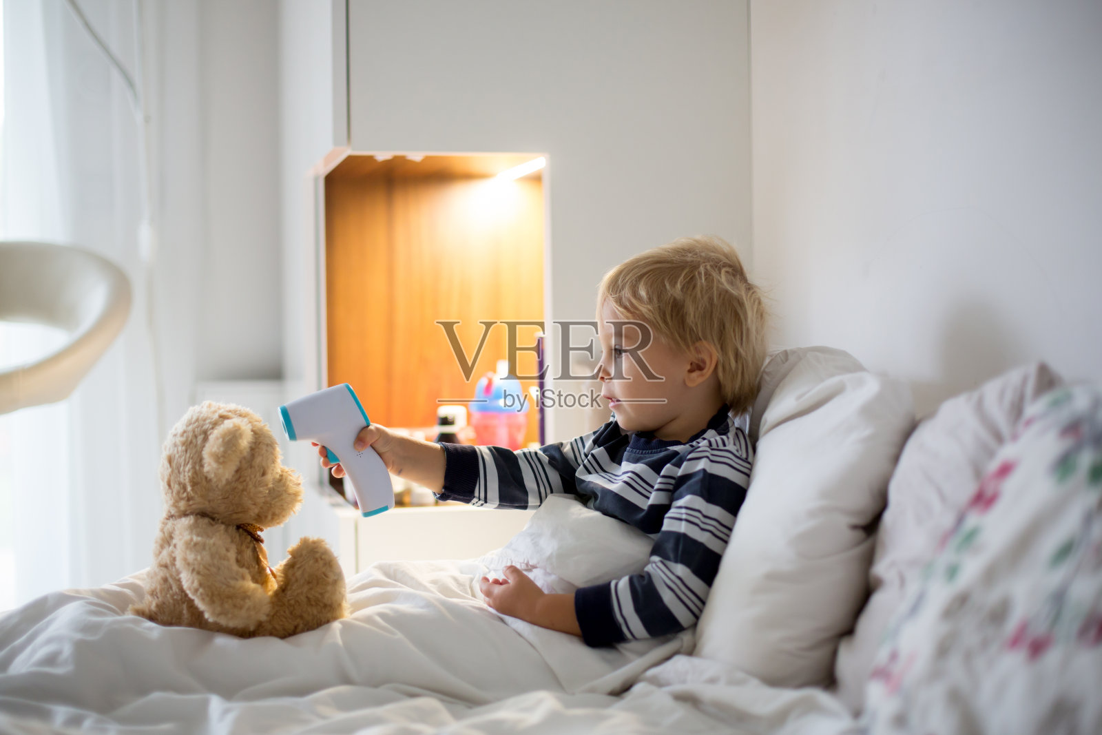 一个蹒跚学步的金发小孩，生病时在床上玩泰迪熊，检查他的体温照片摄影图片