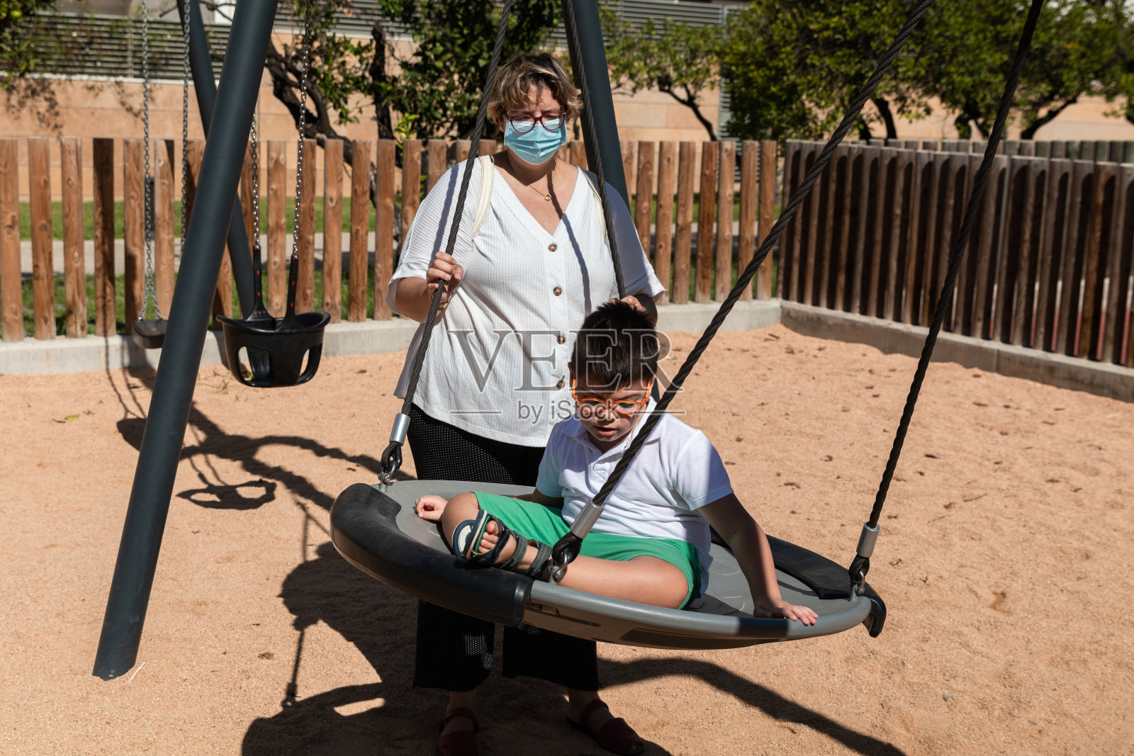 残疾儿童在秋千上，母亲戴着冠状病毒防护口罩照片摄影图片