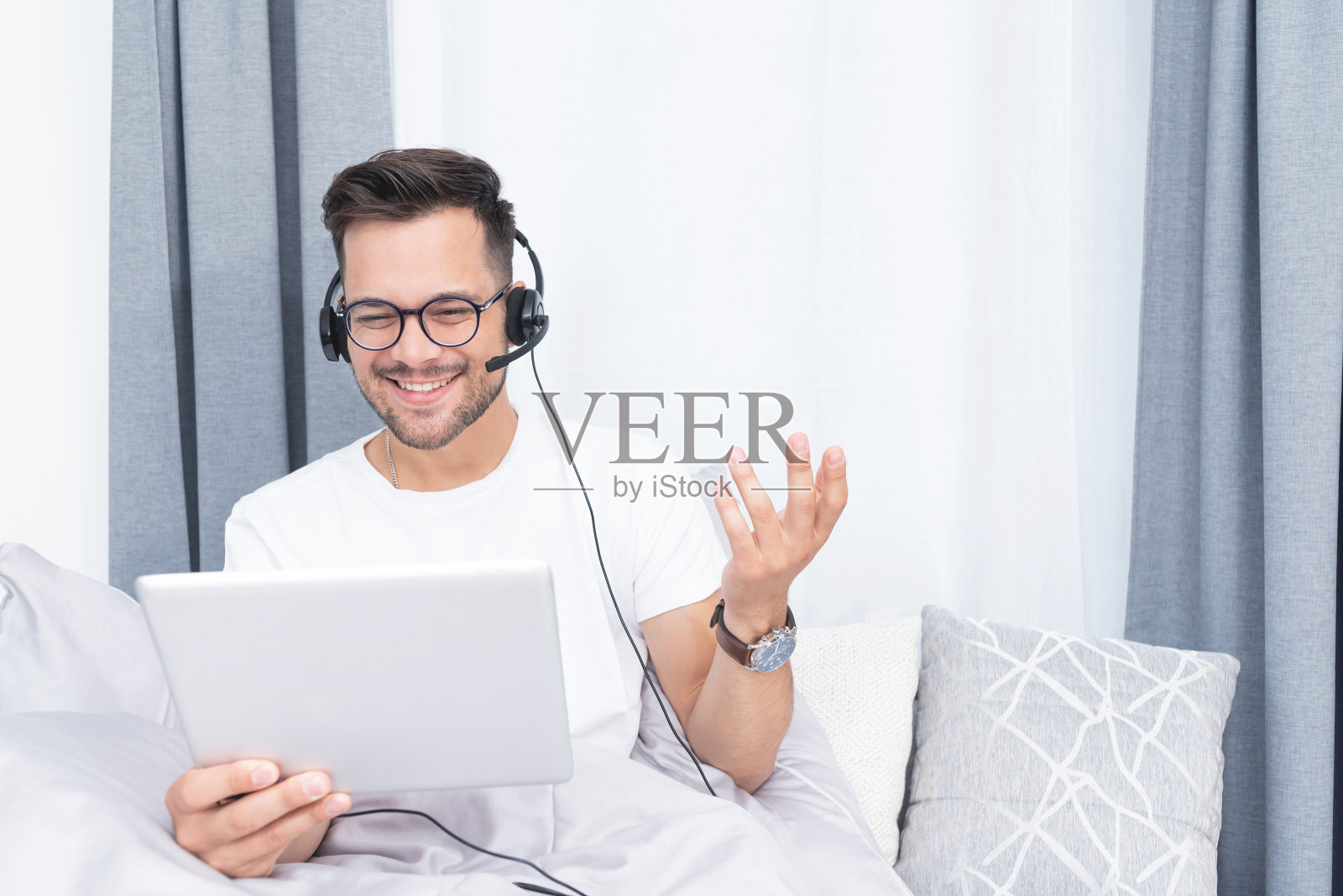 快乐的人使用数字平板电脑和耳机视频聊天，同时放松在床上在家照片摄影图片