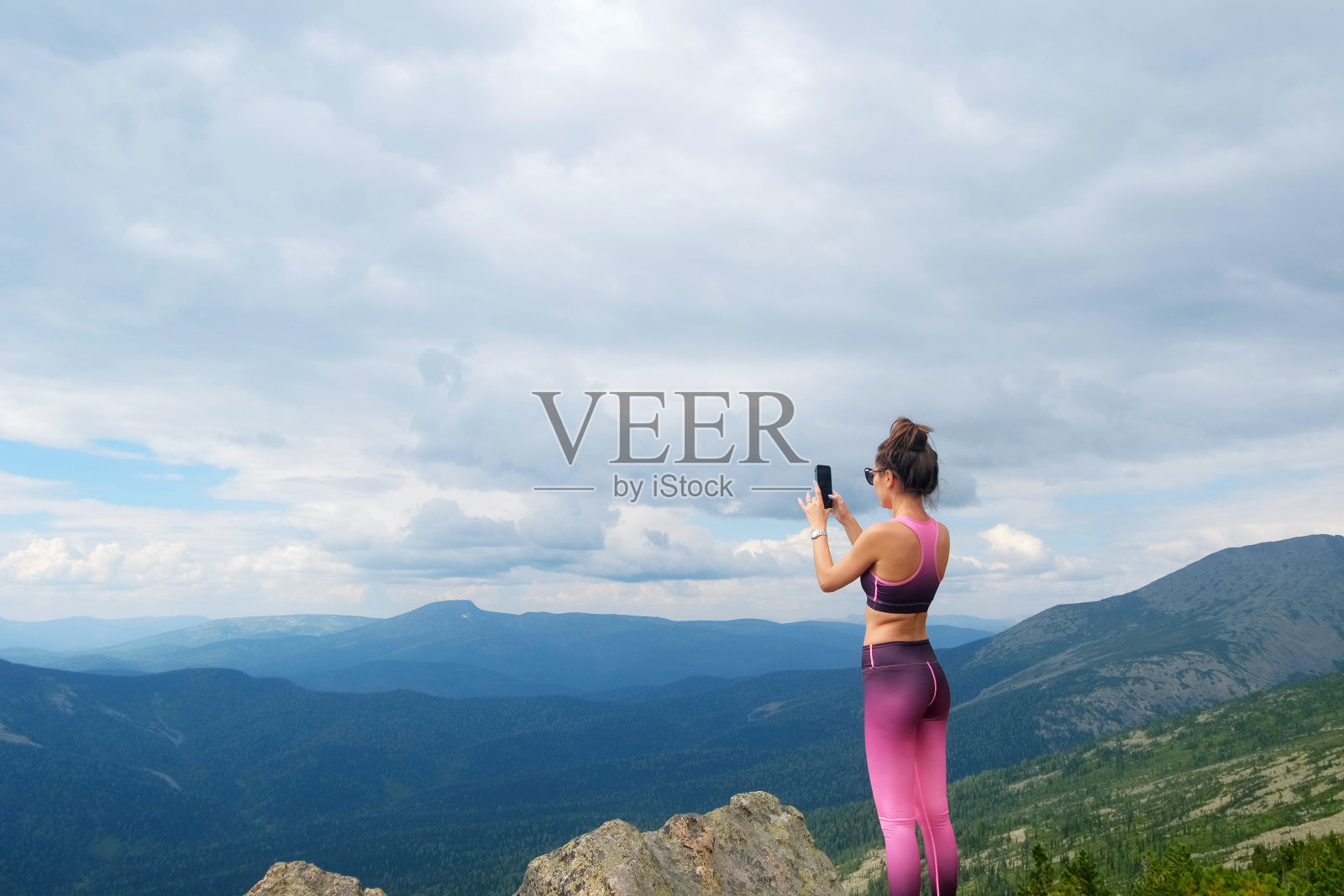 一名女子在山顶的岩石上用智能手机拍照。一名年轻女子在蓝天白云的映衬下爬山。照片摄影图片