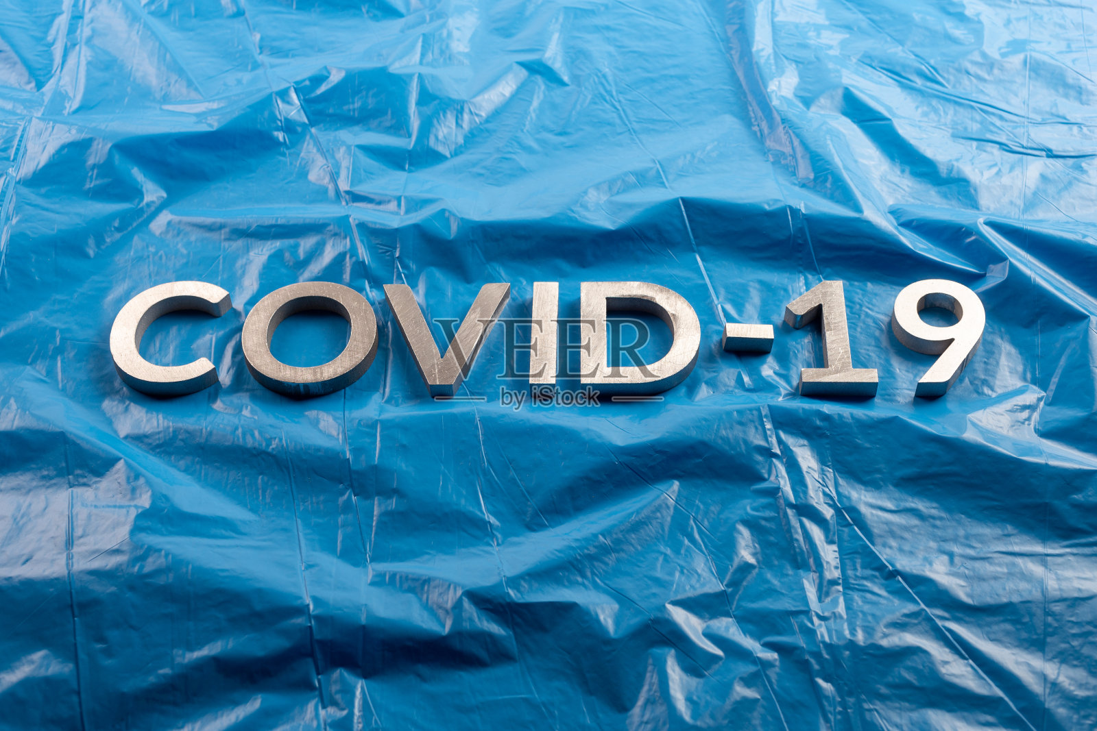 COVID-19这个词是用铝制字母铺在皱巴巴的蓝色塑料薄膜背景上的照片摄影图片