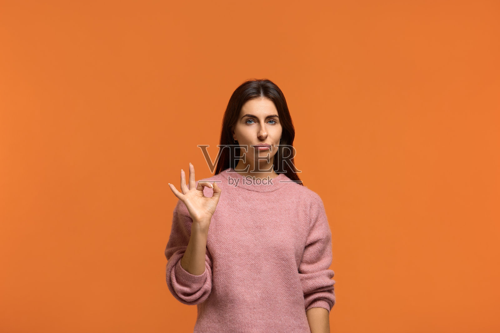 干得好，但我不满意。不坏。漂亮的女人用中性的表情来评判好作品。穿着粉红色针织毛衣孤立在橙色的墙上照片摄影图片