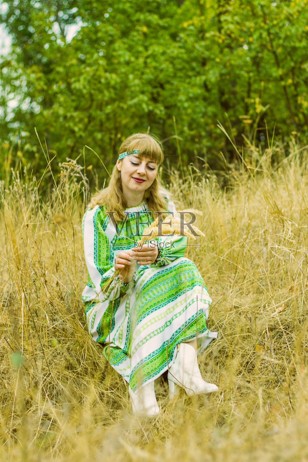 一位穿着绿色民间服装的年轻女子坐在公园的草地上，手里拿着小穗。照片摄影图片