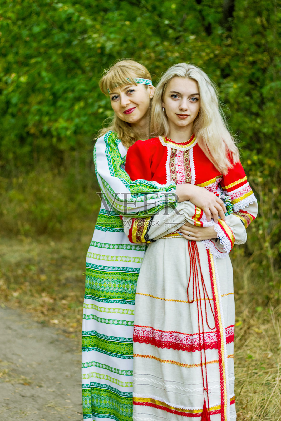 两个年龄不同的年轻女子穿着红绿相间的民间服装，在公园里相互拥抱。照片摄影图片