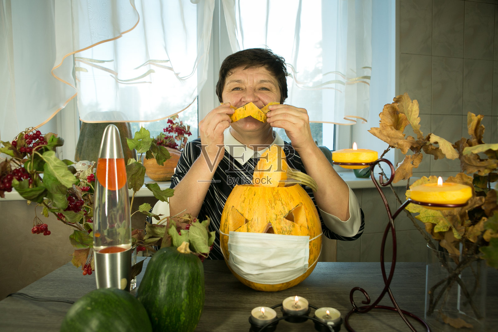 在家里做杰克灯笼。冠状病毒大流行期间的万圣节。一个戴面具的女人站在杰克灯笼前。幽默。照片摄影图片