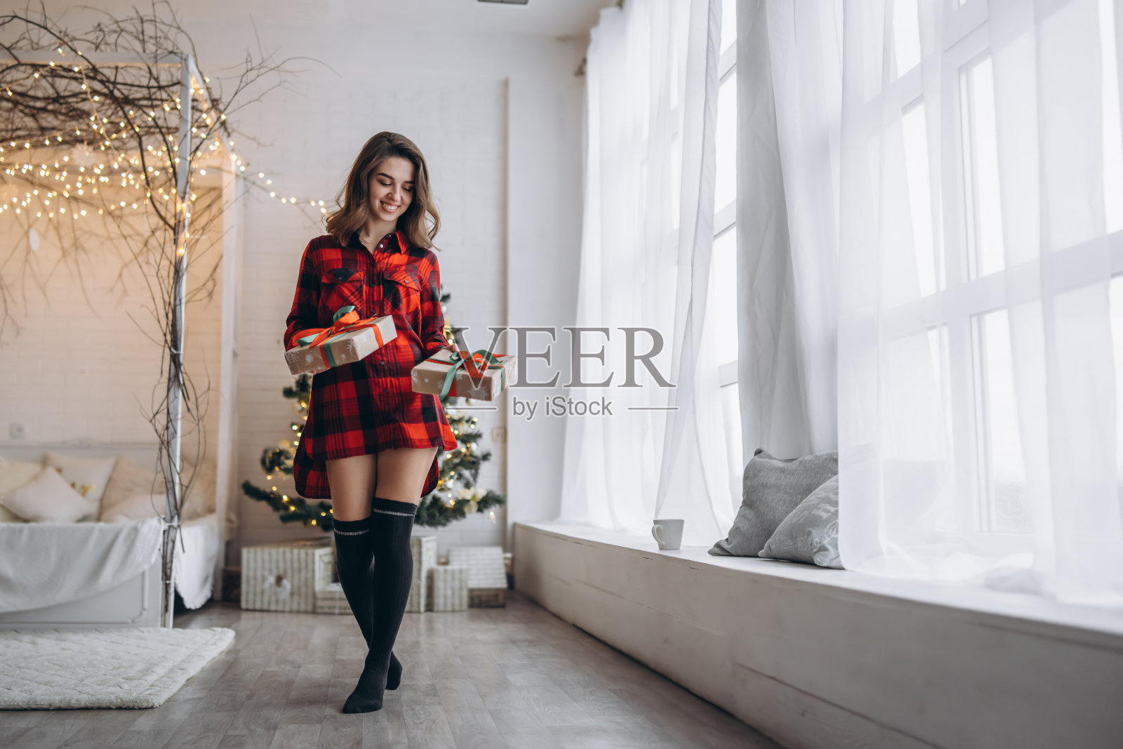 穿着衬衫和袜子的漂亮女人带着圣诞礼盒走在家里，身后是新年树照片摄影图片