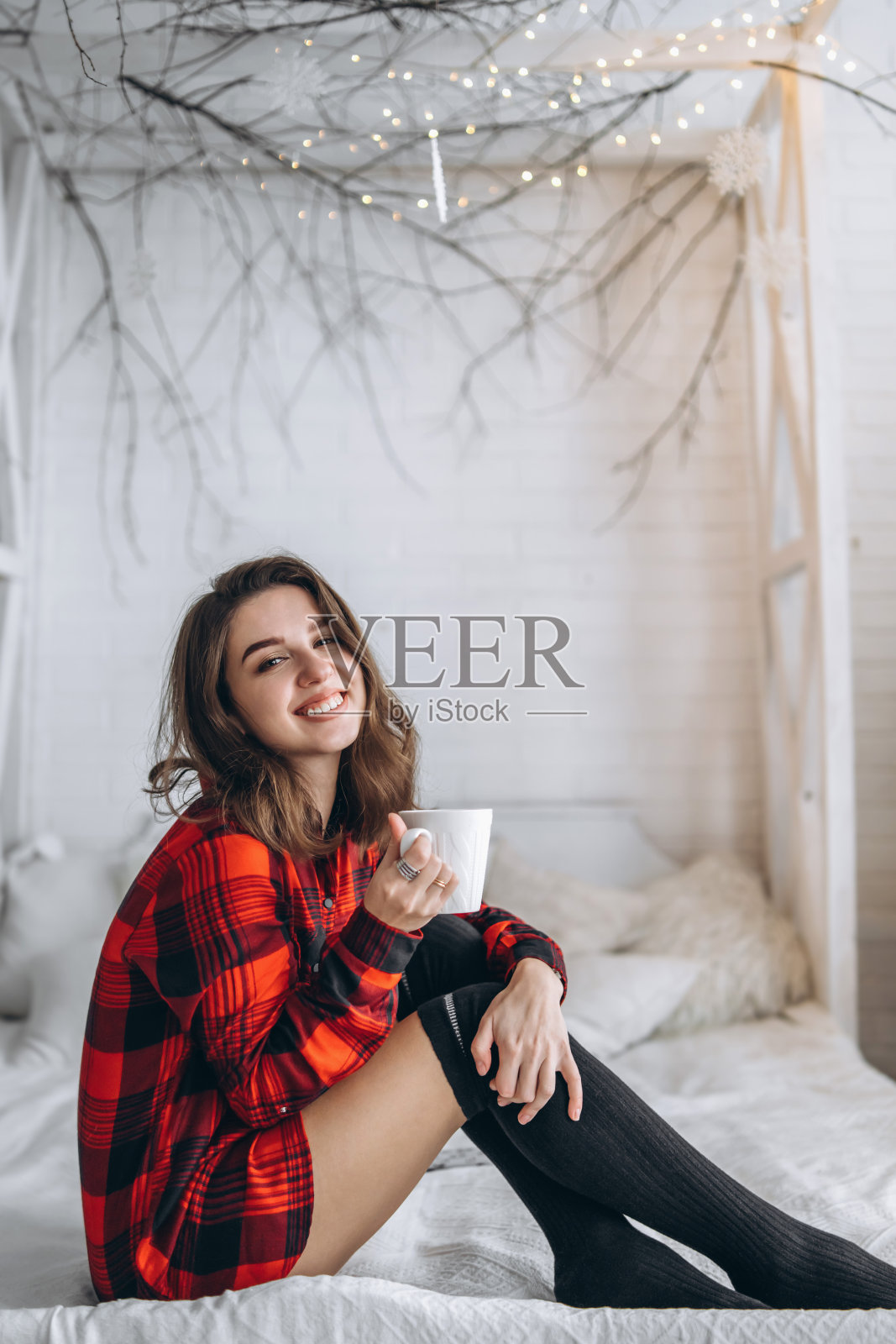 漂亮的黑发女孩躺在床上，穿着红衬衫和温暖的袜子，身后放着一杯咖啡和圣诞彩灯。照片摄影图片